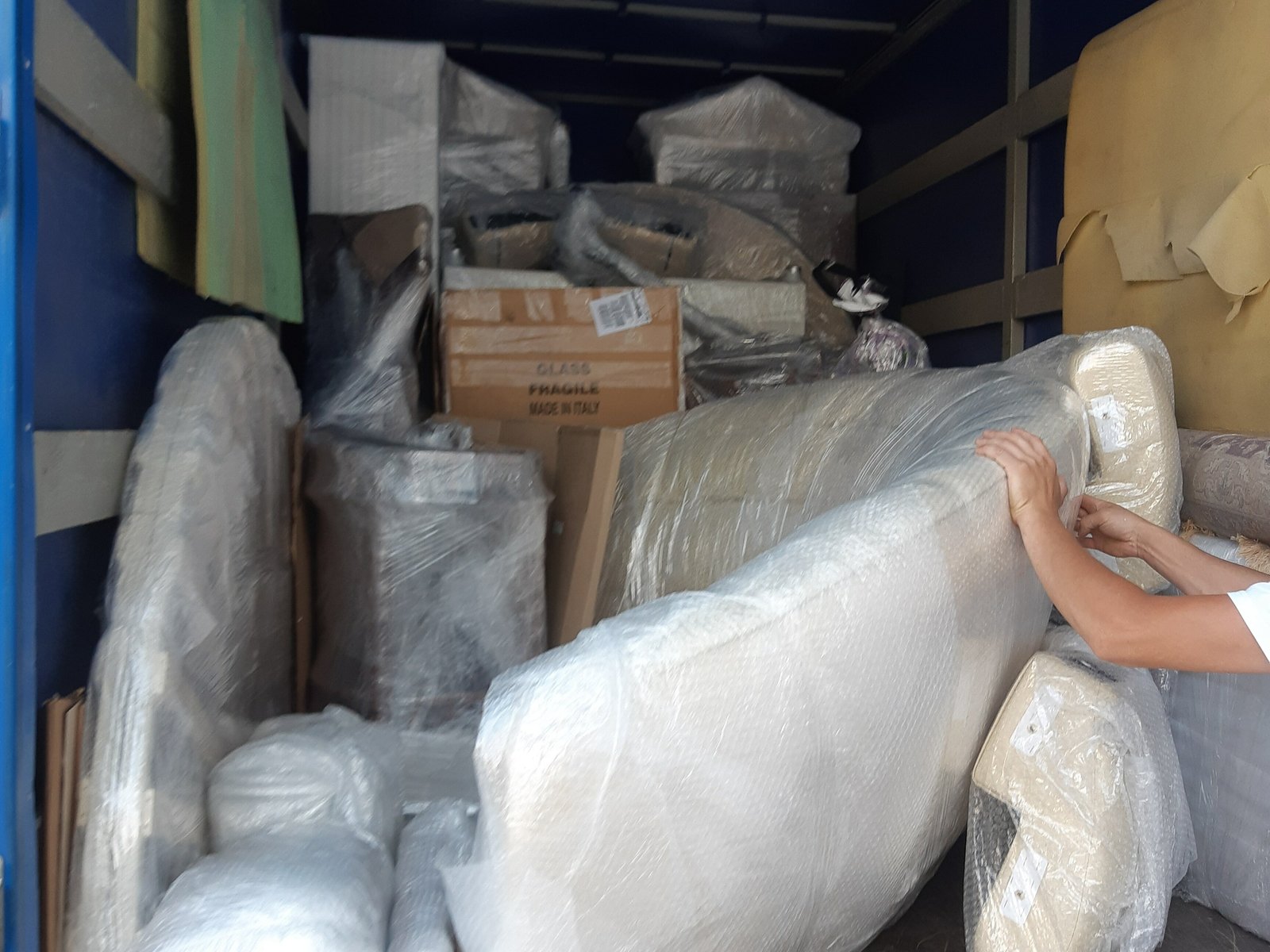 Hands steadying a bubble-wrapped sofa among wrapped furniture inside a moving truck