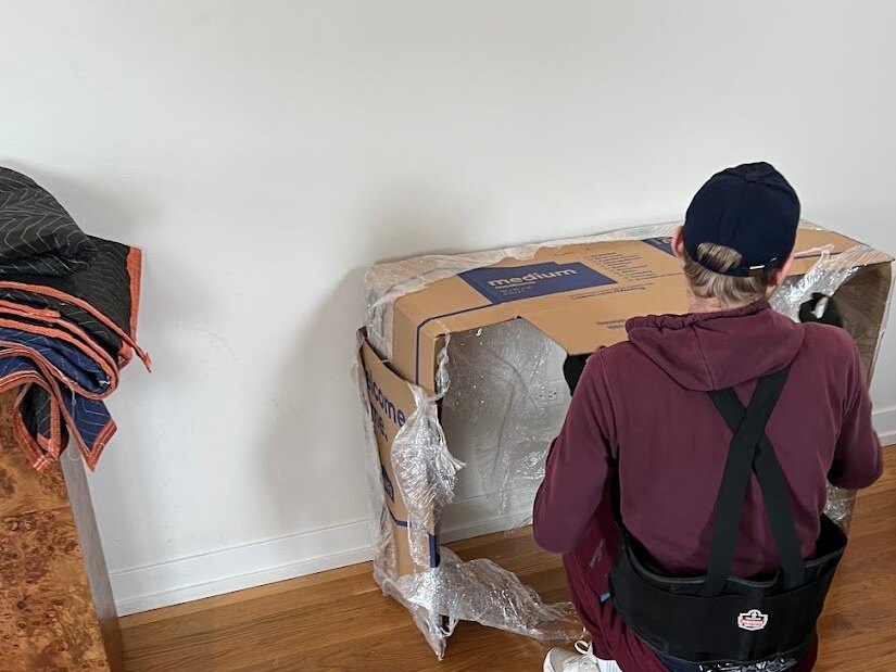 A mover kneeling on a hardwood floor wrapping a piece of furniture in cardboard and plastic film