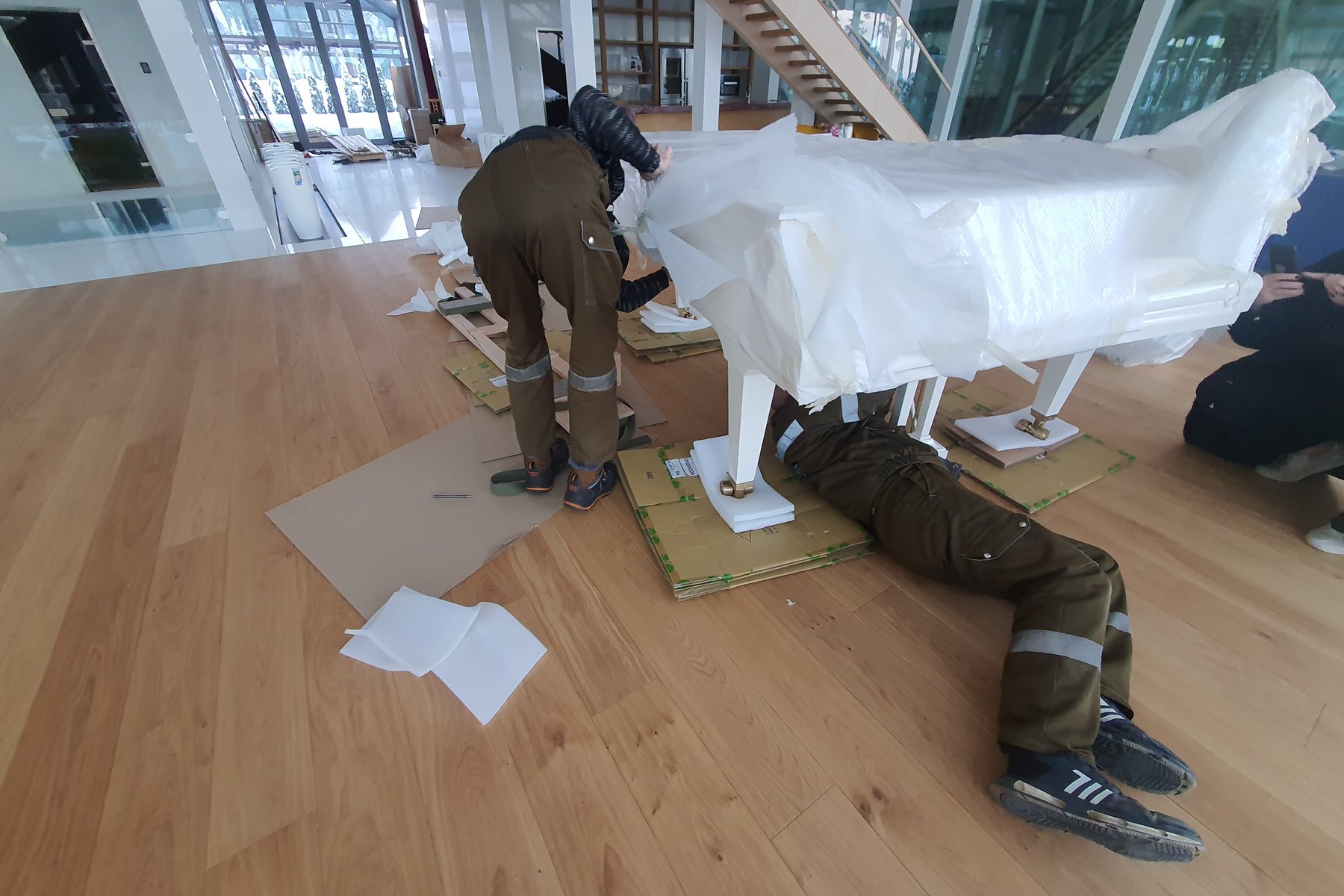 Movers padding and wrapping a white grand piano on a wood floor before transport