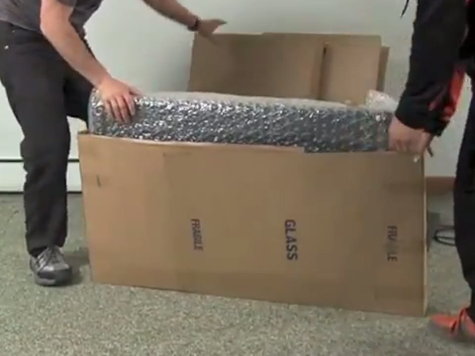 Two movers placing a bubble-wrapped item into a glass carton on a carpeted floor